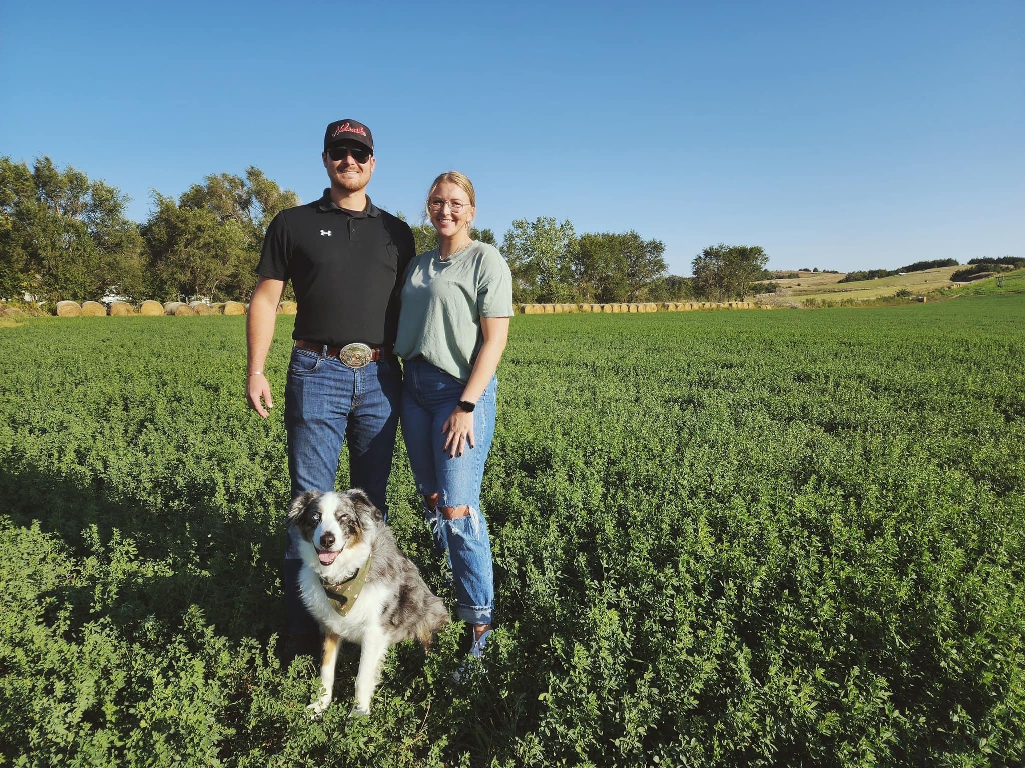 376280835_122098355120048351_5527408383824850832_n A couple stands together in a lush green field with their dog, surrounded by trees and hay bales in the background. The man wears a black shirt and cap, while the woman is in a light green shirt and jeans, enjoying a sunny day on the farm.