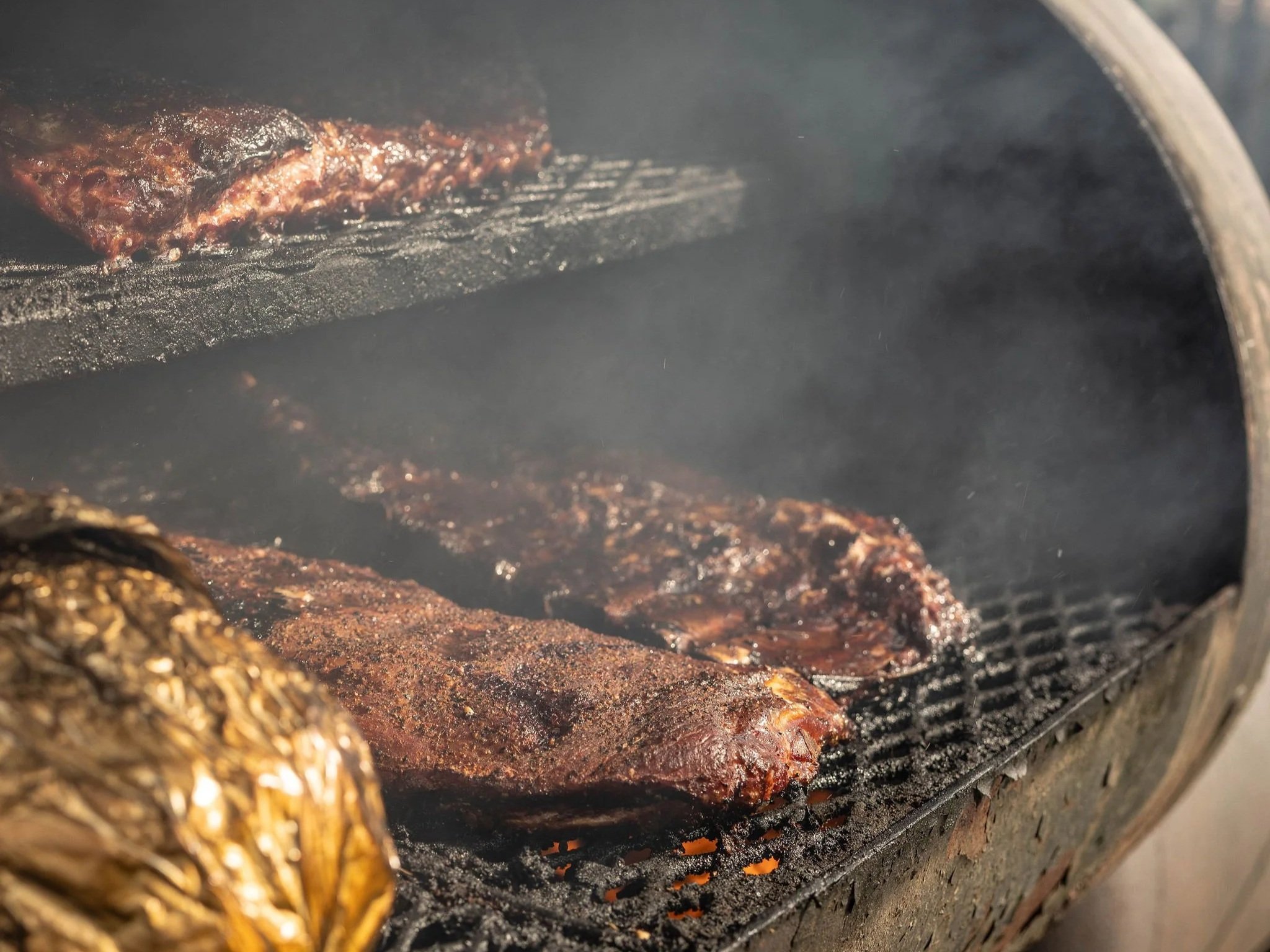 6E3A1969 Smoky barbecue ribs and a wrapped item cooking on a grill, showcasing the rich texture and flavor of smoked meats in a barbecue setting.