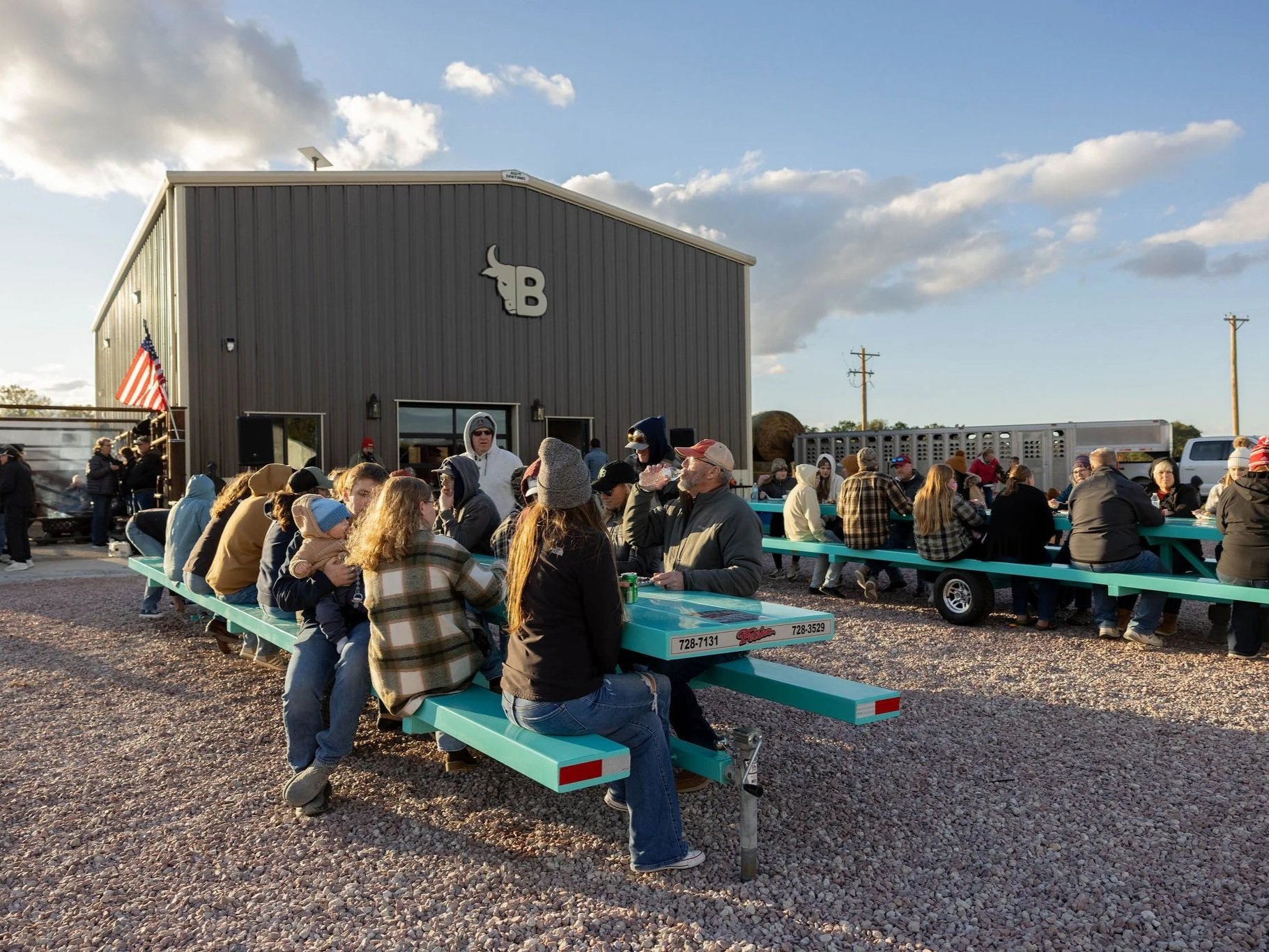 6E3A2823 Outdoor gathering at a rustic venue with people seated at teal picnic tables, enjoying food and drinks. The scene features a modern gray building with a bull logo and an American flag, set against a clear sky, showcasing a vibrant community atmosphere.